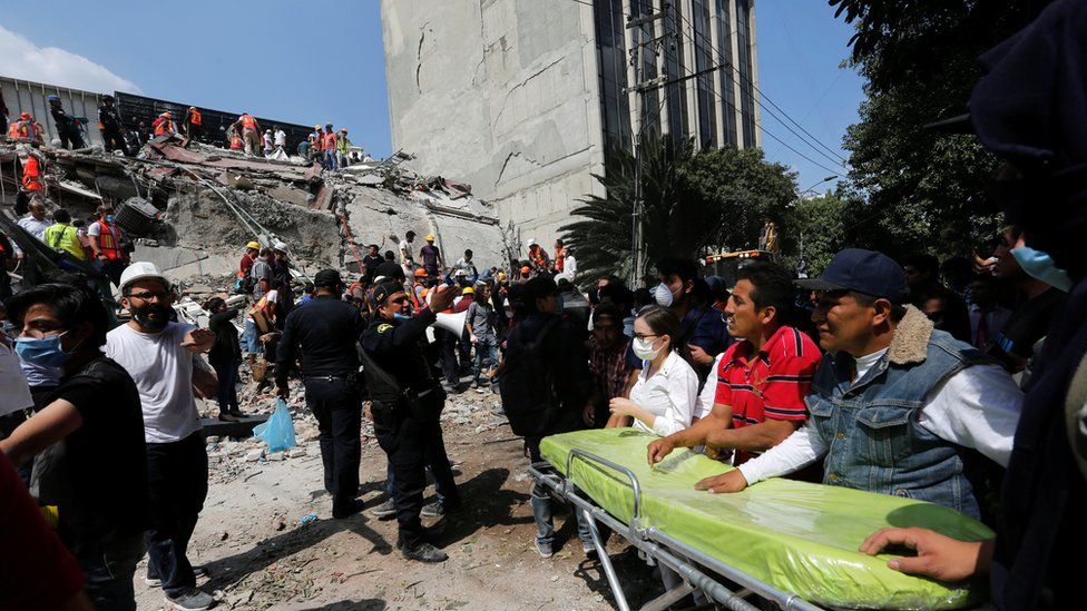 rescuers in front of a collapsed building that is right next to a building still standing, in Mexico City