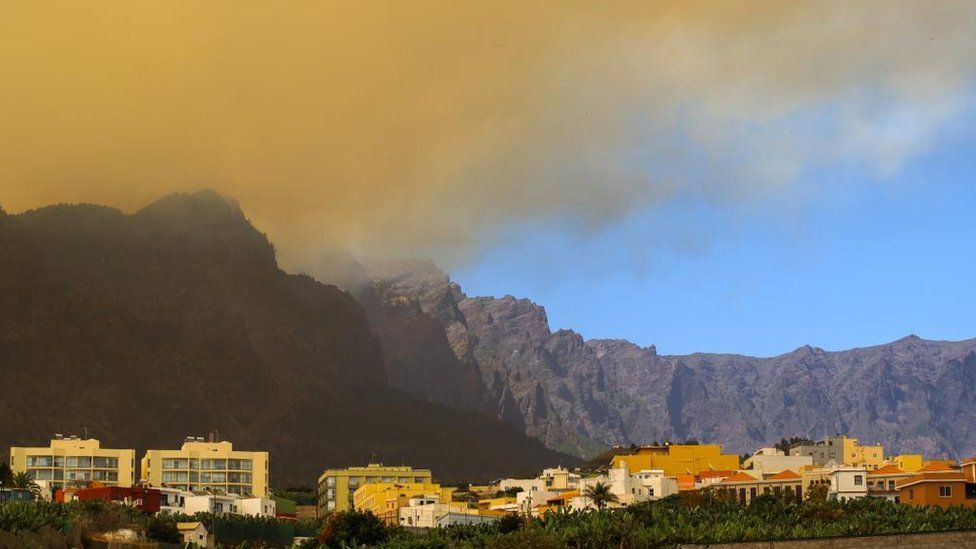 Smoke billows from a hill during a forest fire on the Canary island of La Palma. A row of buildings is visible in the foreground.