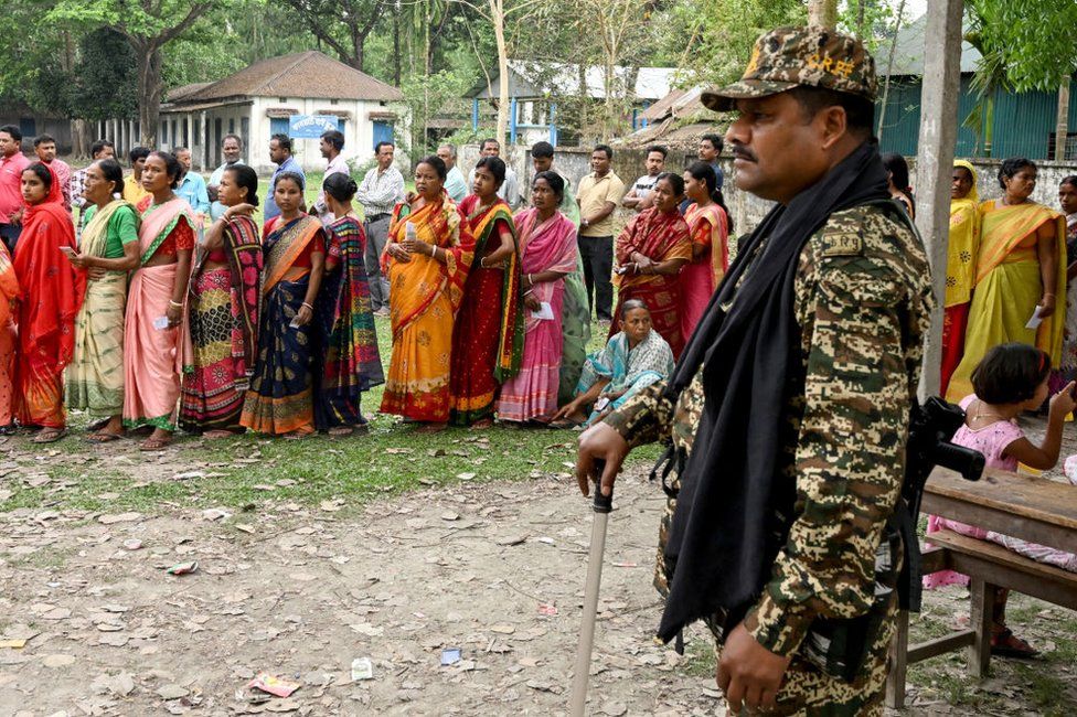 A security personnel stands guard beside people in a queue waiting to cast their votes as voting starts in the first phase of India's general election at a polling station in Kalamati village, Dinhata distict of Cooch Behar in the country's West Bengal state on April 19, 2024