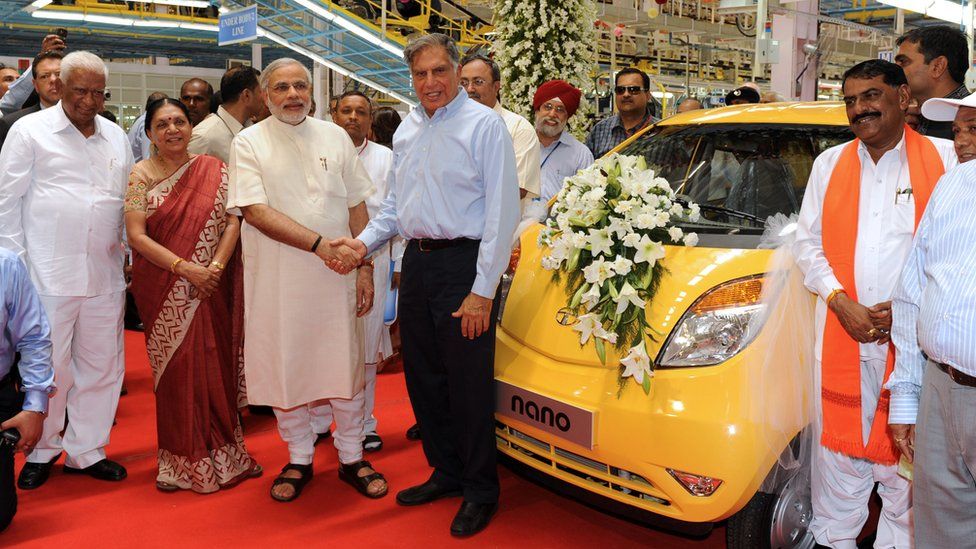 Narendra Modi, then governor of Gujarat, shakes hands with the chair of Tata group at a new factory. A yellow car is adorned with white flowers.