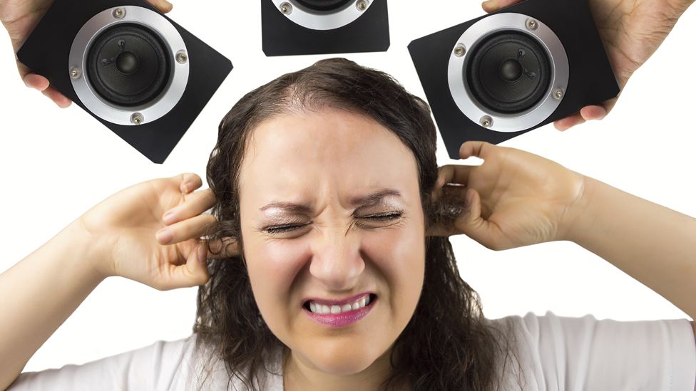 Woman holding her fingers in her ears, surrounded by speakers