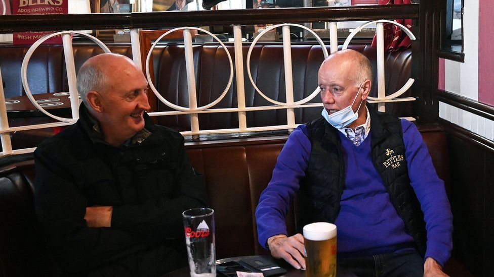 Two older men, both bald, sitting indoors in a bar with drinks in front of them. The man on the right is wearing a mask he has pulled down below his chin.