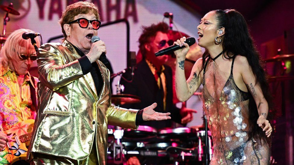 Elton John performs with Rina Sawayama, with Elton on the left and Rina on the right. Elton is singing into a microphone while wearing a golden blazer with black shirt, and red tinted glasses. Rina is wearing a iridiscent sequin outfit, facing Elton and singing into the microphone, with an ear piece visible in her left ear. The background has Elton John's band, with instruments visible.