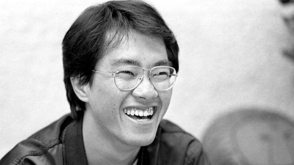 Akira Toriyama, pictured in black and white. Akira is a Japanese man with dark floppy hair. He grins, looking to the right of the camera, his eyes crinkled in the corners. He wears metal framed round glasses and a black bomber jacket.