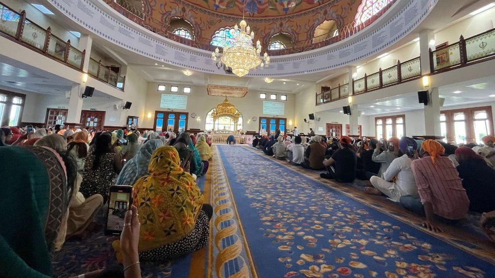 A busy Gurdwara with many worshippers inside. Viewing the congregation from the back of the room, a range of colourful head coverings can be seen on the seated worshippers. In the distance a lone figure kneels in prayer in front of an altar. A lavish chandelier hangs from a domed ceiling painted with an ornate pattern.