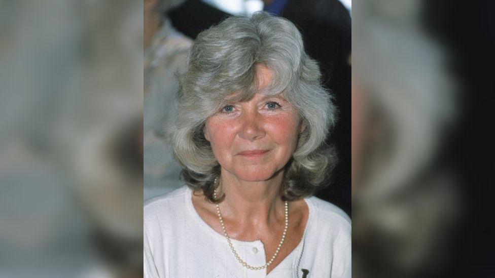 Jilly Cooper wearing a white top and pearl necklace smiling at the camera