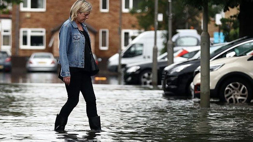 A woman in wellies walks through floodwaters in a carpark near houses in Battersea, London