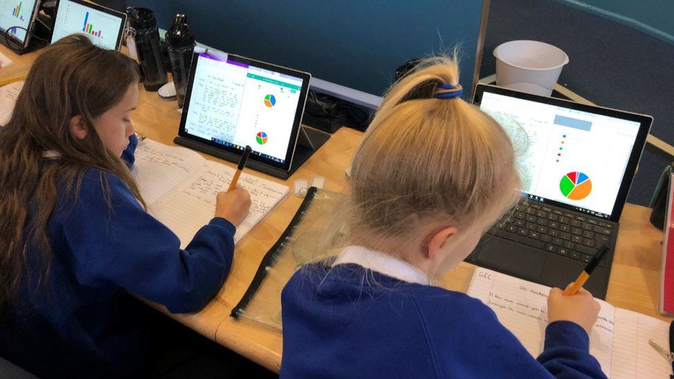 Two girls at a primary school in Exeter writing in school exercise books, with laptop screens open in front of them