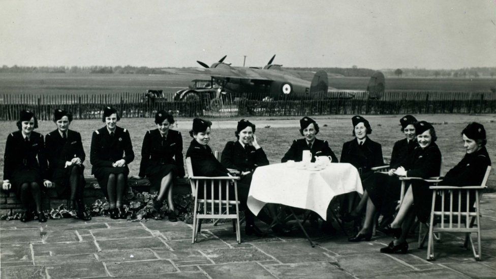 A black and white photo of an airfield where 10 women in uniform and hats are sitting around a table in front of an RAF De Haviland Flamingo aircraft