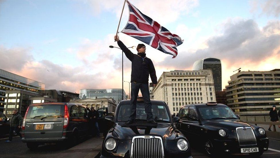 A black cab driver waves a Union Jack flag whilst standing on a taxi.