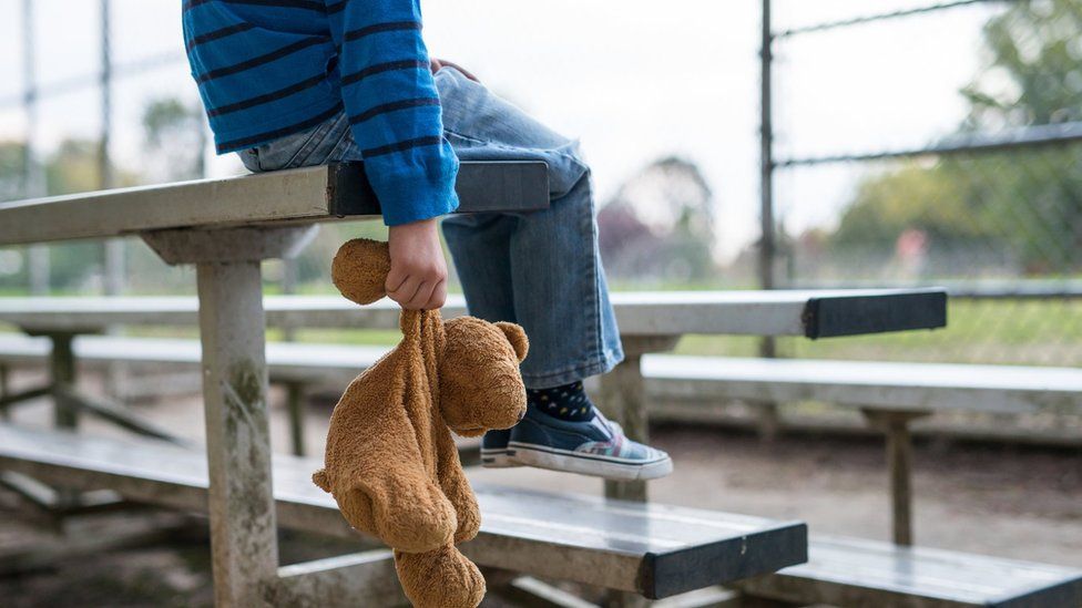 A young boy sitting by himself on a stand of benches, holding a teddy bear