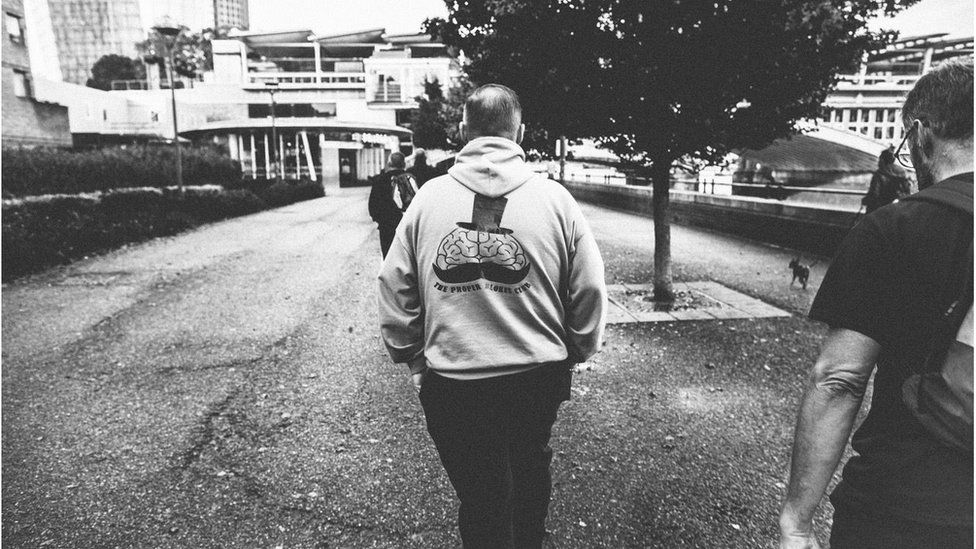 A man wearing a hoodie with the The Proper Blokes Club logo walks with his back to the camera