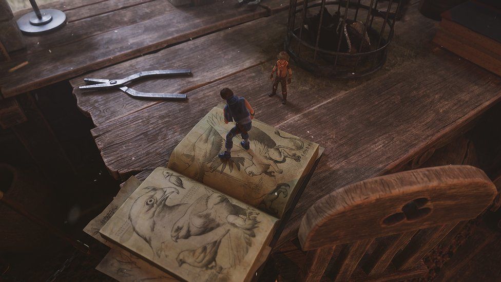 A game screenshot of a boy with black hair standing on a giant book, open to a page featuring a sketch of a mythical, bird-like creature with a lion's tale. To his right a younger boy stands on a table surface, close to a pair of silver pliers that, like the book, is far bigger than both human characters. To the right of the shot we can see the top of a massive chair, suggesting the boys are in the home of a giant.