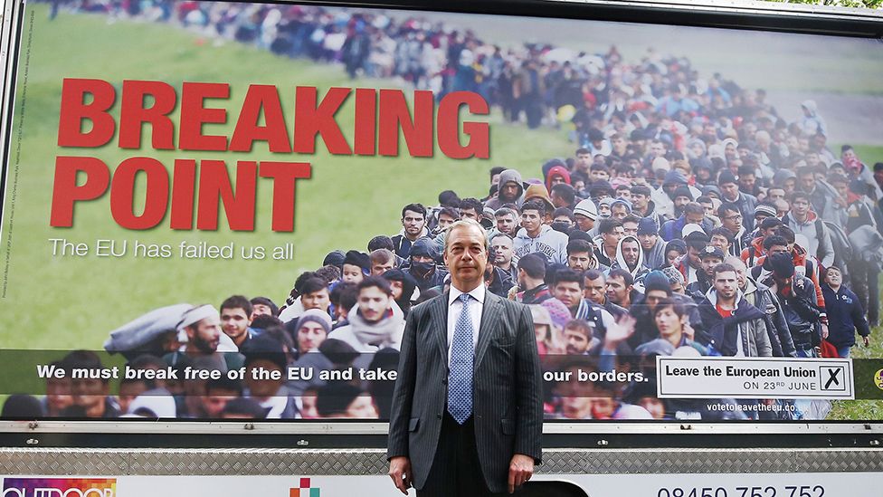 A man in a grey suit, white shirt and light blue necktie stands in front of a billboard with a photo showing a long line of people presumed to be asylum seekers or migrants. The queue snakes off into the distance. In red, block capitals the words "Breaking Point" appear on the left of the picture. Underneath that it says "The EU has lied to us all". At the bottom, white text on a black bar reads "We must break free of the EU and take back control of our borders". In the bottom right a white rectangle bears the text "Leave the European Union on 23rd June" and there is a black box with a black cross in it, suggesting a mark on a ballot paper.