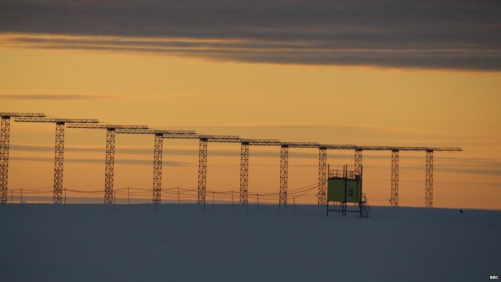 A row of tall pylons in front of a pale yellow sky. There is a small hut in the midground