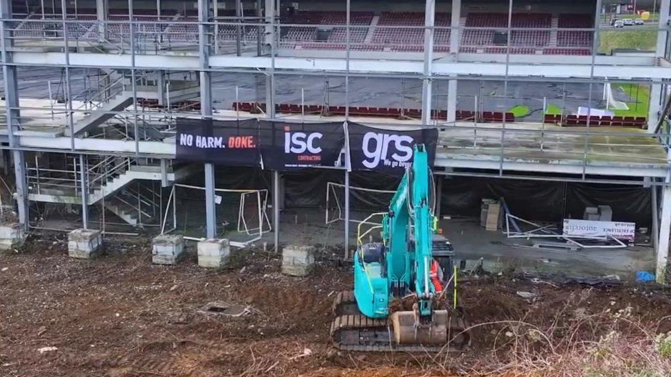 Blue/green digger picking up soil behind the partially-completed East Stand