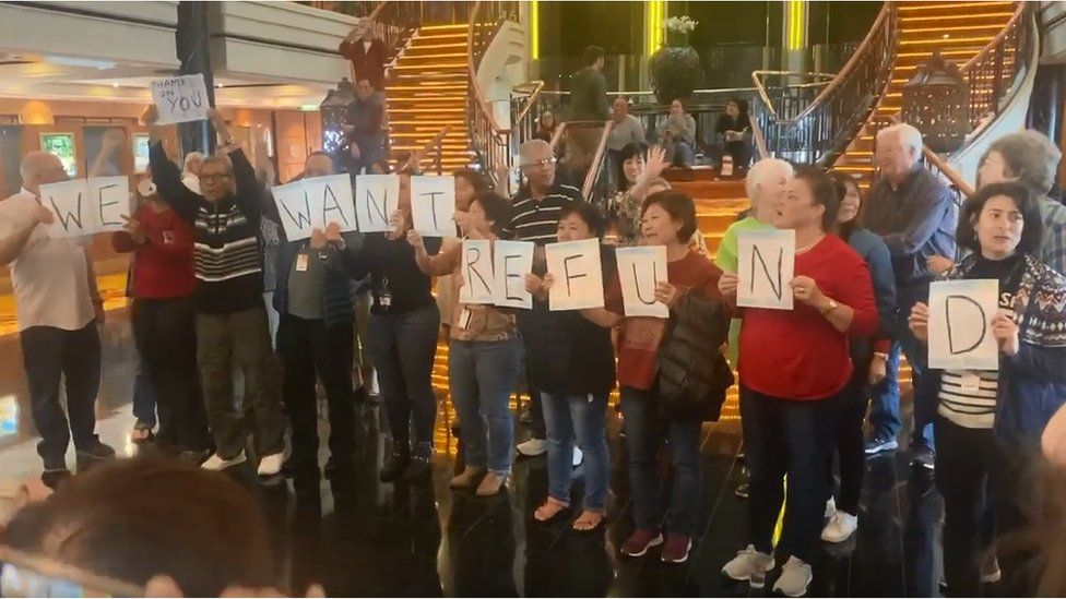 Passengers crowd hold signs spelling out "we want refund"