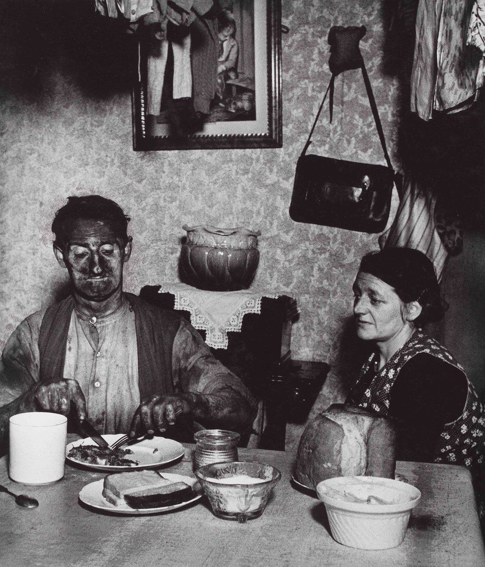 A photograph of a miner eating a meal at a dinner table as a woman looks on