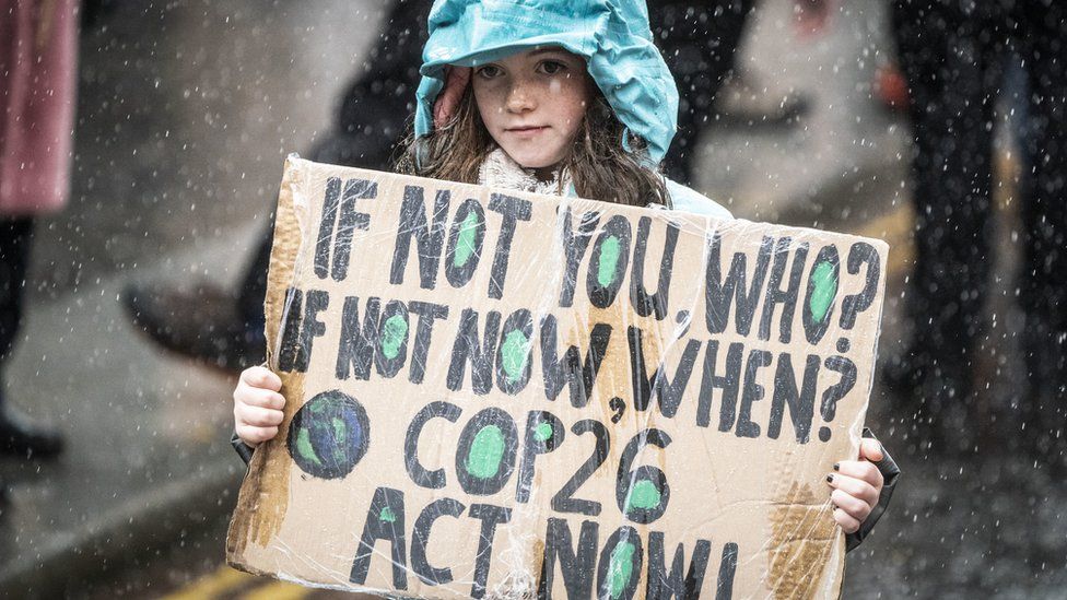 Девушка держит табличку с надписью «Если не ты, то кто? Если не сейчас, когда? COP26: Действуй сейчас»