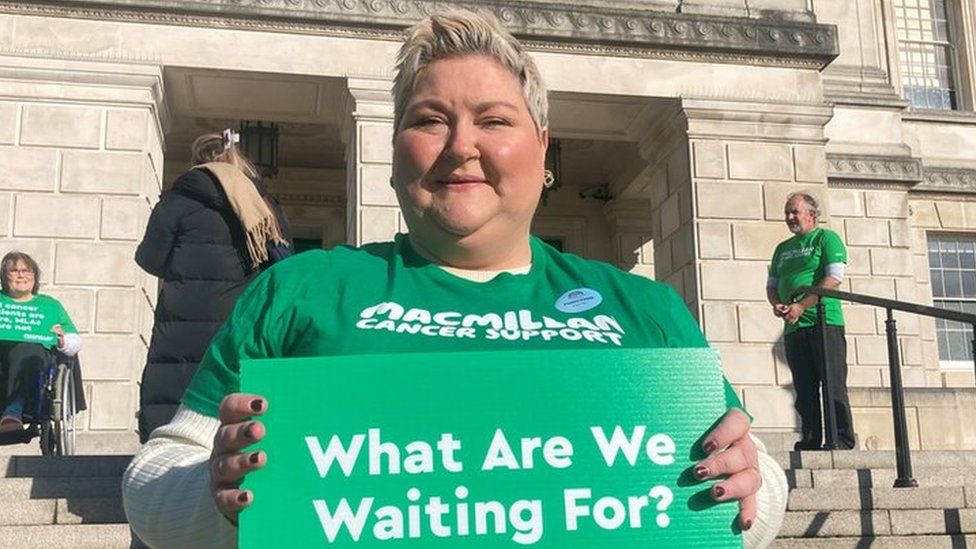 Colleen McCallion standing in front of Stormont holding a sign reading "What are we waiting for?"