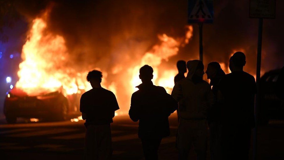 People are silhouetted against the flames after a large number of cars were set on fire on Ramels vag in Rosengard, Malmo