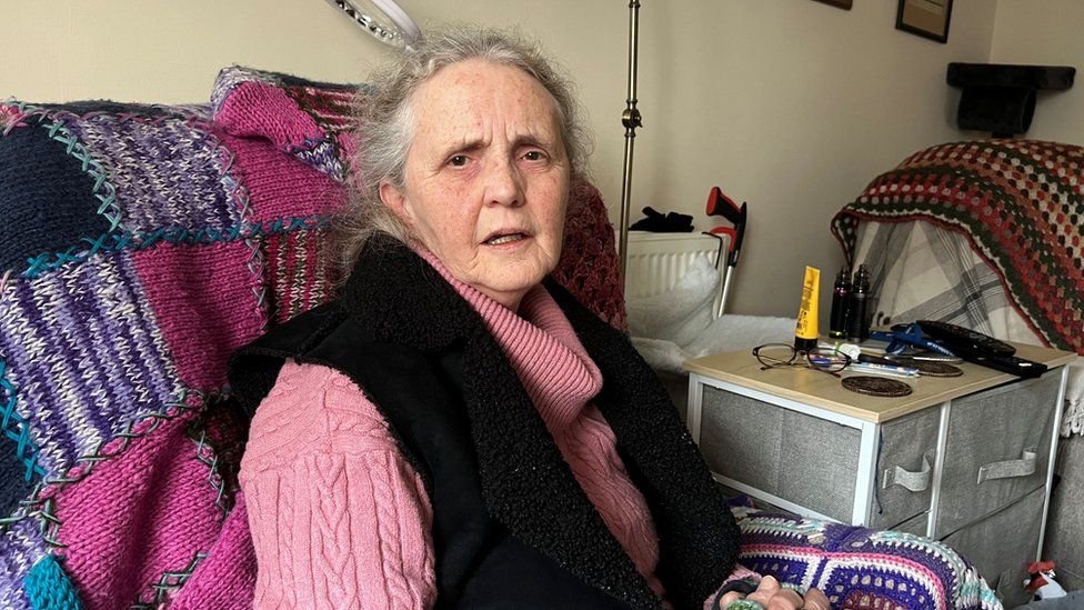 Karen Mitchell, an elderly woman in a pink jumper, sitting on a chair with a patchwork blanket over it