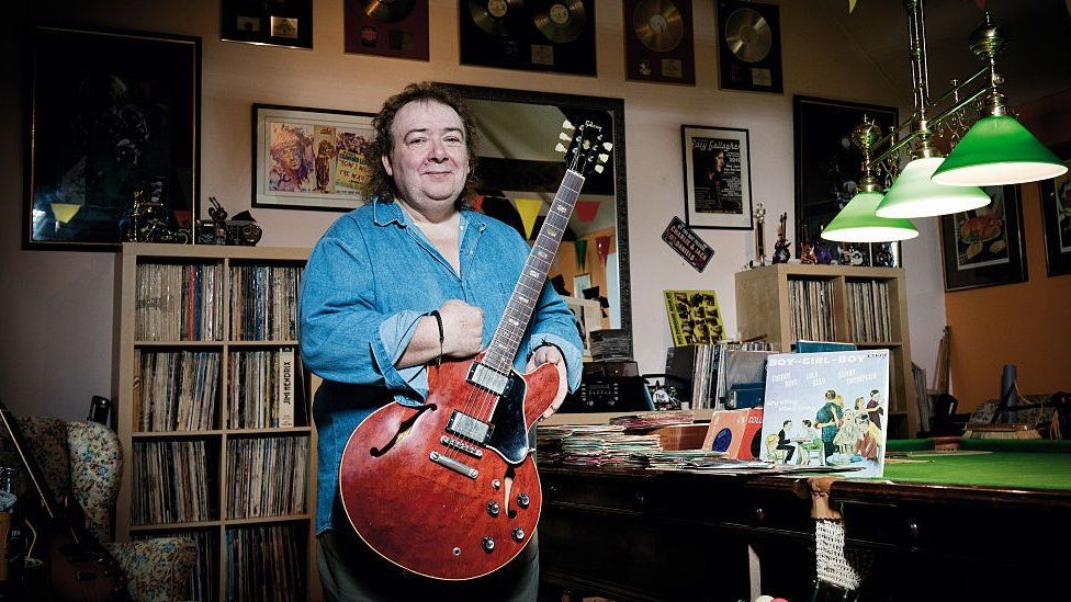 Bernie Marsden holding a guitar standing next to a snooker table covered with 45s single records