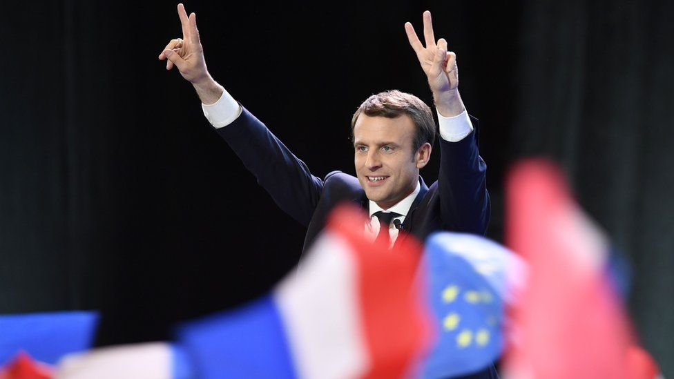Emmanuel Macron hold his hands up in a "peace sign" gesture with the French and EU flags visible in the foreground