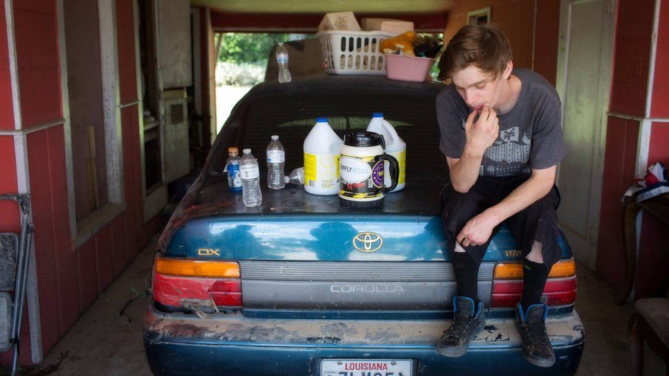 Jesse Poche, 23, sits on one of his automobiles in the carport of his home as 20 some odd volunteers assisting him with demo are swirling around. As he searched for some of his belongings he was able to preserve in plastic bins, he said "slowly but surely, I will regain everything I lost". Denham Springs, LA.