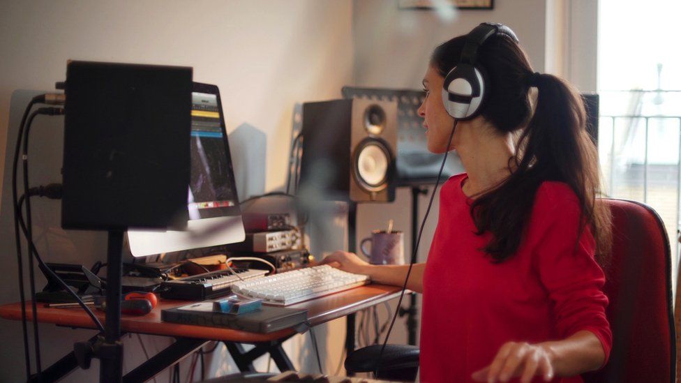 A woman with headphones on and surrounded by music equipment sits at her computer