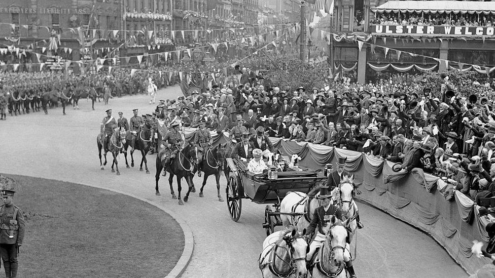 Large crowds of people watch as King George V and Queen Mary travel in a horse-drawn carriage to Belfast City Hall to open the new Northern Ireland Parliament