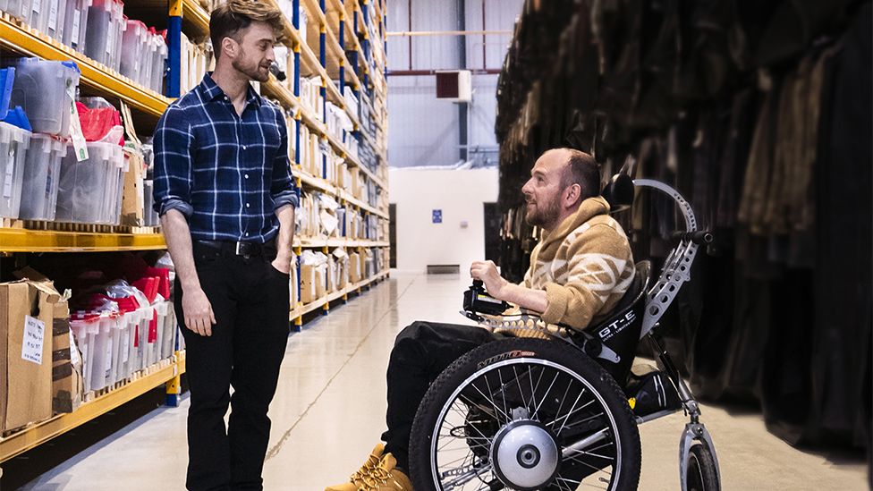 Daniel Radcliffe and David Holmes. Daniel is standing wearing a blue checked shirt with white lines and black trousers. David is in a wheelchair with black wheels and silver rims, wearing a brown and white top looking up at Daniel. The background is of a warehouse with lots of shelves stacked with items in plastic boxes.