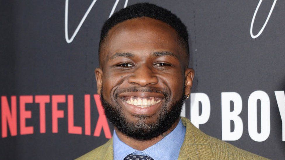 Joshua Blissett attends the World Premiere of "Top Boy 2" in London in 2022. Joshua, a black man in his 20s, wears a pale blue shirt under a light green blazer. He has short hair and a trimmed beard and is smiling at the camera.