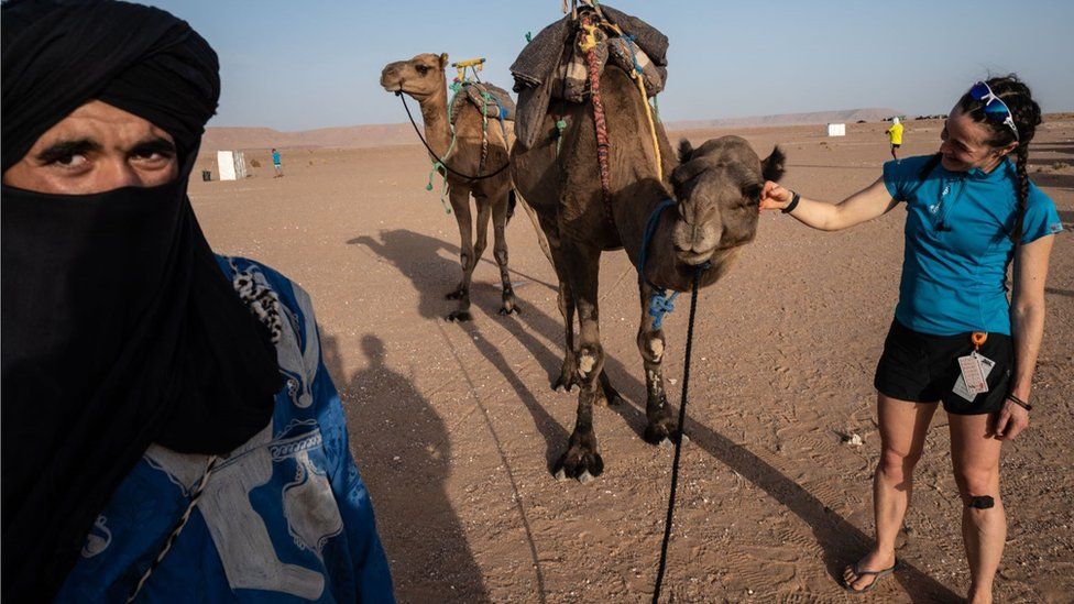 A woman competitor of the Marathon des Sables in the Sahara desert in Morocco is stroking a camel. There is a vast space of sand behind her. A man with a black turban is also in the shot.