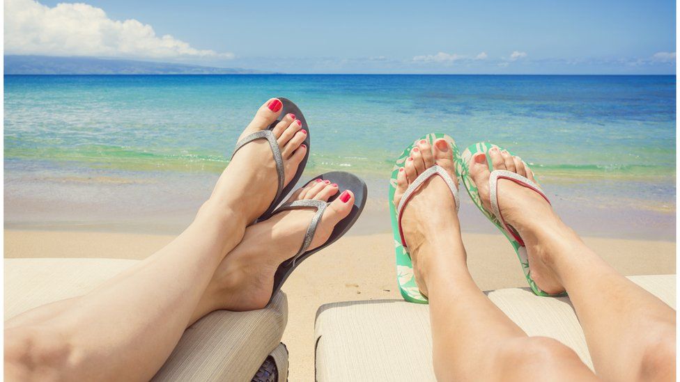 Two women lie next to one another on sun loungers wearing flip flops with the ocean in the background