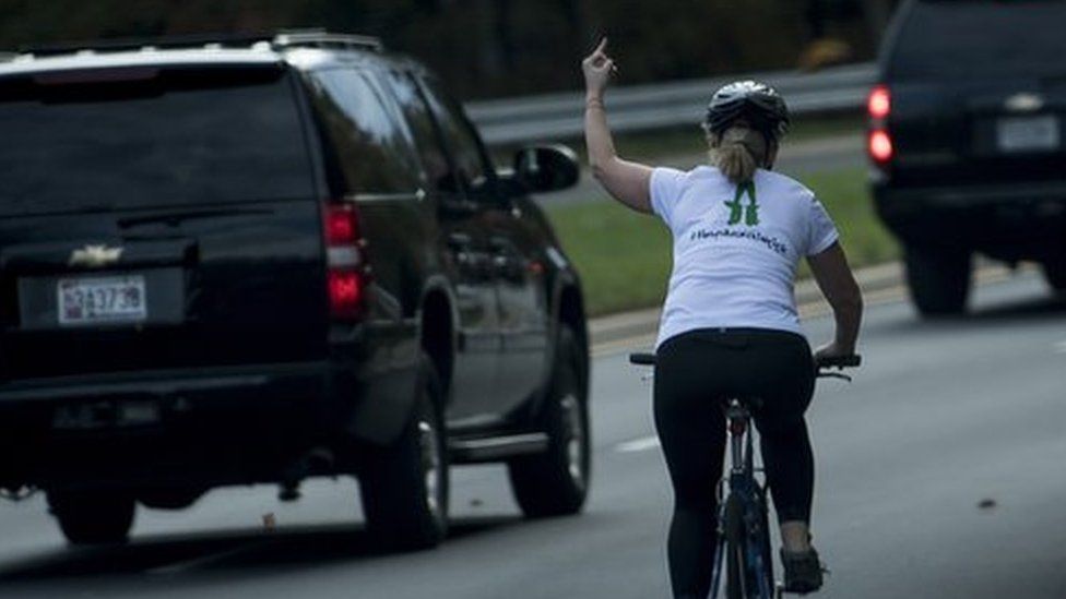A line of motorcade cars and Juli Briskman on a cycle making a middle finger gesture