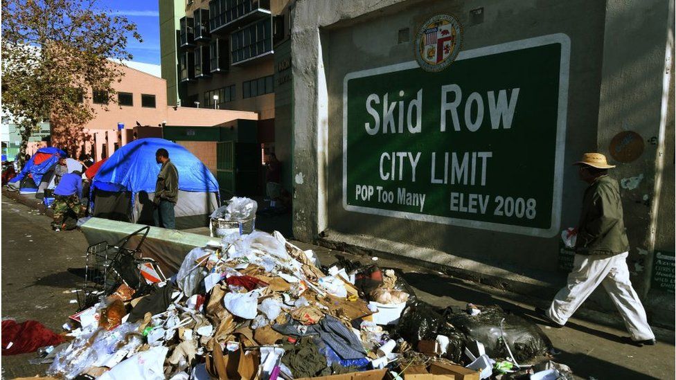 Trash and tents in front of a mural reading 'Skid Row, population too many, elevation 2008'