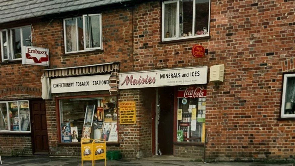 Old-fashioned shop named "Maisies" in brick building with adverts for cigarettes and ice cream by the front door