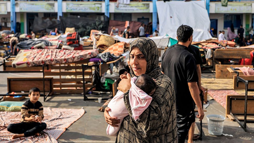 A woman walks carrying an infant in the playground of a school run by the United Nations Relief and Works Agency for Palestine Refugees (UNRWA) agency that has been converted into a shelter for displaced Palestinians in Khan Yunis in the southern Gaza Strip on October 25, 2023