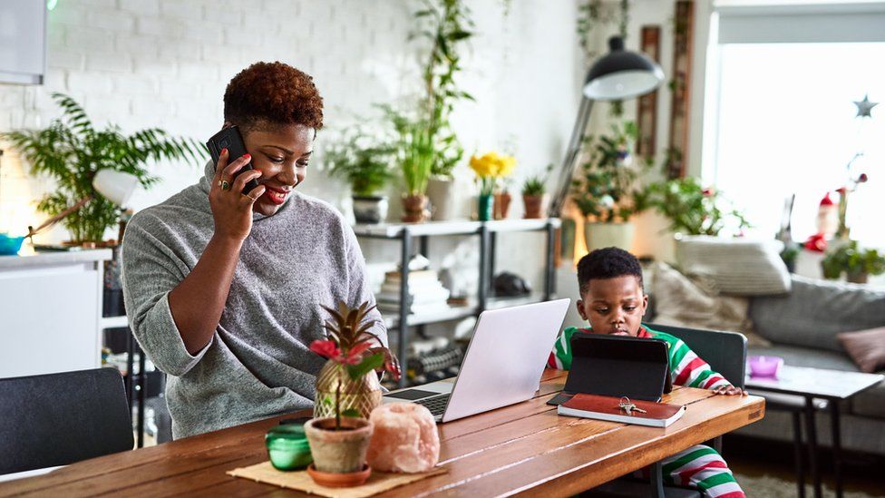 A woman on on the phone with a laptop in front of her. A young boy sits near her, looking at a tablet.