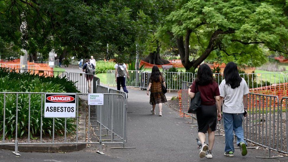 People walk past an asbestos sign that is displayed at Victoria Park in Chippendale