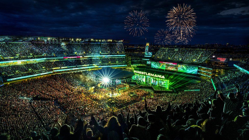 A wide shot of the crowd and the stage of Wrestlemania on Saturday night. In the background of the stage, there are fireworks being set off in the night sky during Night One of WrestleMania 40 at Lincoln Financial Field.
