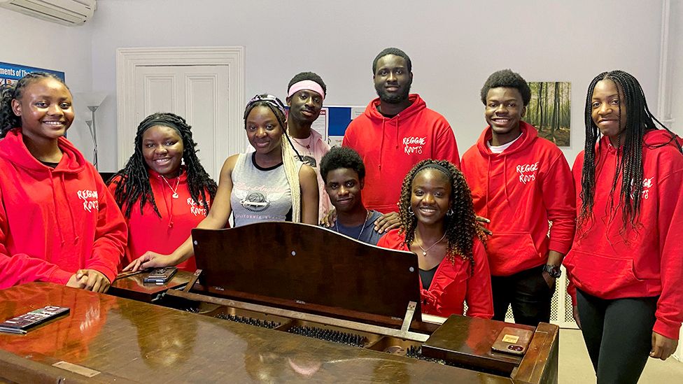 Девять членов хора Reggae Roots собрались, улыбаясь в камеру, вокруг пианино в репетиционной комнате Большинство из них носят красные толстовки с напечатанным на них названием группы.