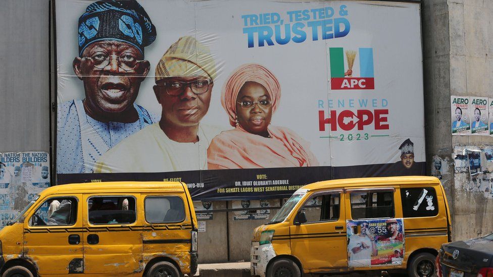 Two yellow buses in front of a campaign poster of Tinubu and the Lagos state governor