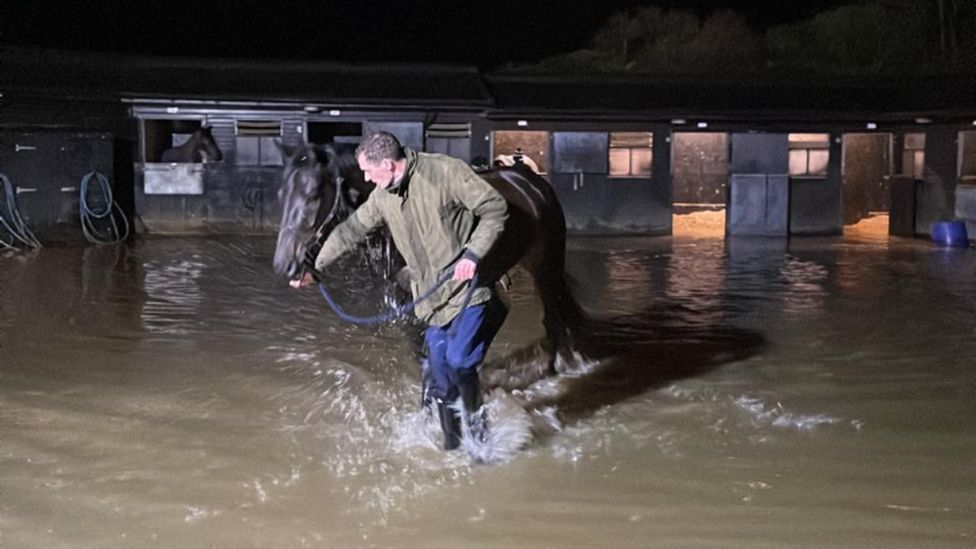 A man leading a horse away through a few inches of flood water at a stable yard