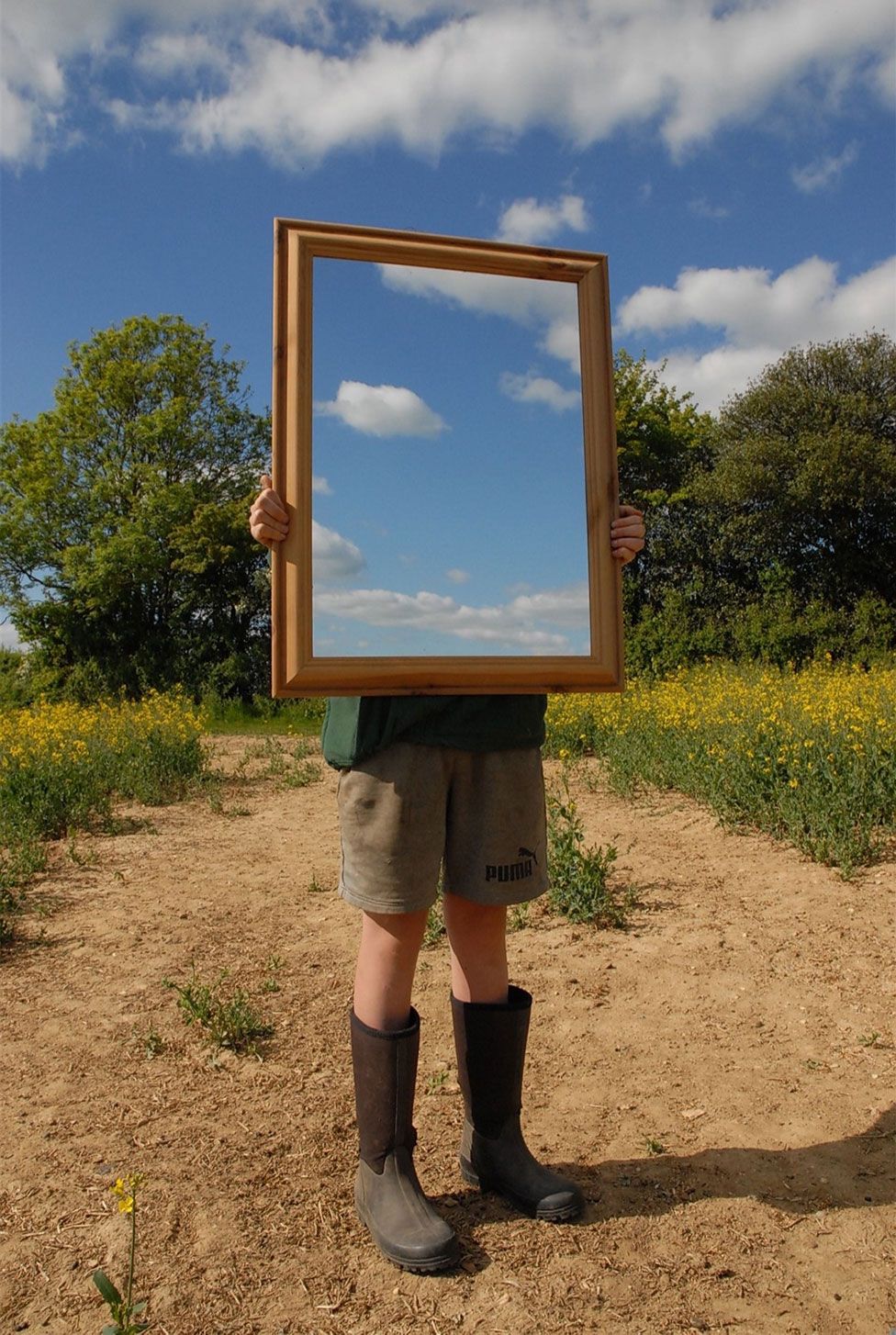 A person stands in a field and holds a mirror showing the sky