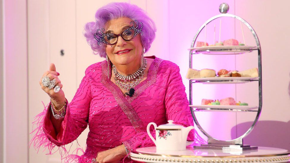 Dame Edna sits near a high tea stand