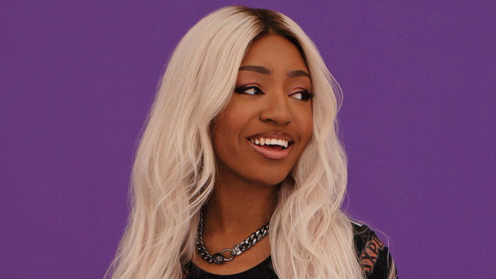 A young, black woman with blonde hair stands in front of a deep purple plan background. She looks off to the side, smiling, her long hair spilling past her shoulders. She's wearing a chunky silver chain, both ends of are linked together by two fairly large hoops than join just under her throat.