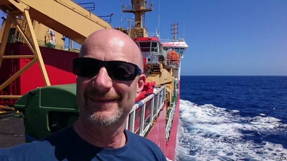 Peter Gibbs in foreground onboard the ship. Bright blue sky and deep blue ocean is behind him.