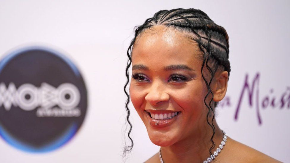 Yinka Bokinni at the 2024 Mobo Awards in Sheffield. Yinka is a 34-year-old black woman. She wears her hair braided and tied back. She smiles, looking to the left of the camera and wears a diamond necklace.